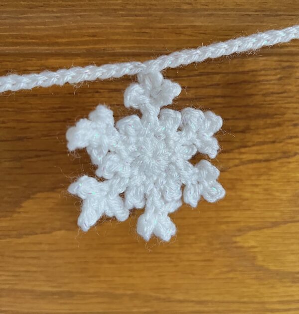 A hand crocheted garland of 10 snowflakes made using a white acrylic yarn with a shimmering thread running through. Approximate length 121cm.