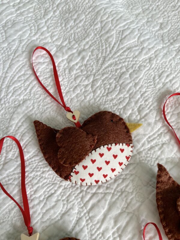 A hand cut and hand stitched, felt and fabric robin, with a red ribbon hanging loop and wooden button detail. Robins approximate size 8cm w x 5cm h. (excluding hanging ribbon.)