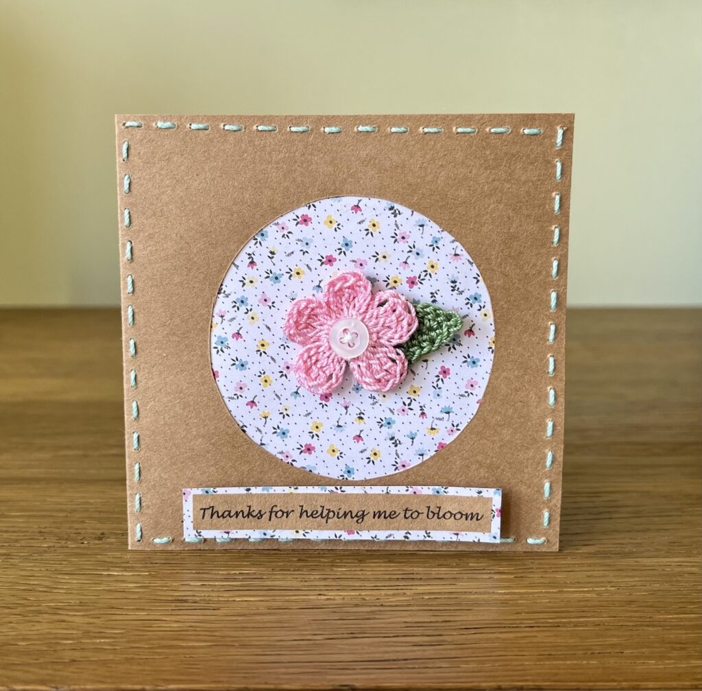 A single crocheted pink flower with button detail and a green leaf, with hand sewn boarder and Thanks for helping me to bloom message on the front. This card has a blank white paper insert for you to write your own message. Handmade, using 100% cotton.