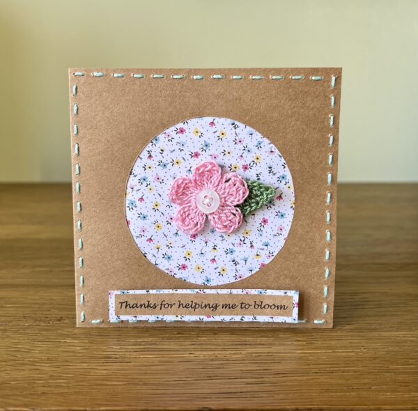 A single crocheted pink flower with button detail and a green leaf, with hand sewn boarder and Thanks for helping me to bloom message on the front. This card has a blank white paper insert for you to write your own message. Handmade, using 100% cotton.