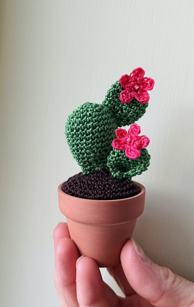A handmade, crocheted mini green cactus with bright pink flowers that have a glass bead centre. The cactus is sat in a terracotta pot.