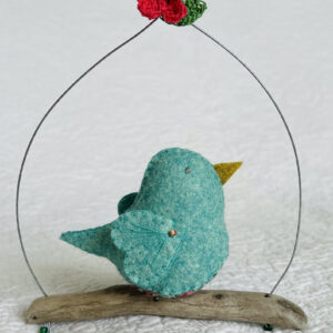 A single medium sized bird, handmade in green felt with a cotton leaf and berry print fabric chest and hand embroidered detail on the wings. This bird is sat on a natural driftwood perch with a wire hanger that is decorated with a pink flower and green leaf detail.