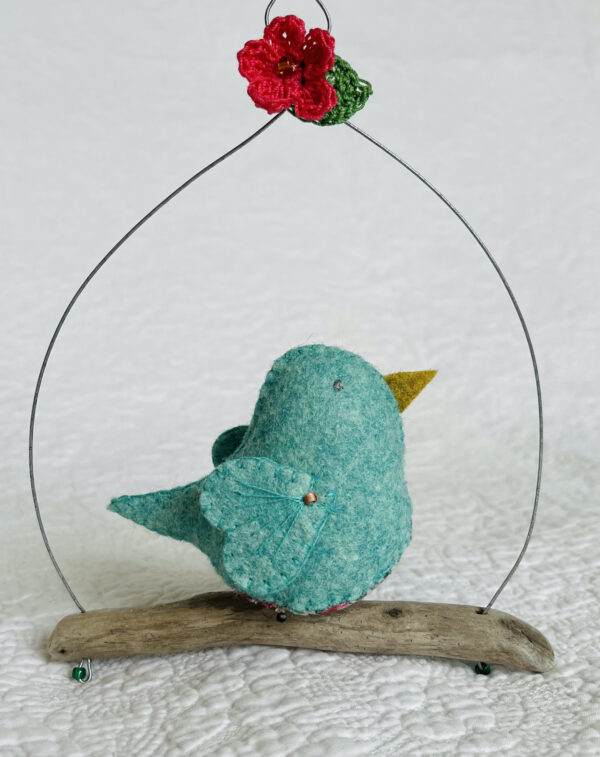 A single medium sized bird, handmade in green felt with a cotton leaf and berry print fabric chest and hand embroidered detail on the wings. This bird is sat on a natural driftwood perch with a wire hanger that is decorated with a pink flower and green leaf detail.