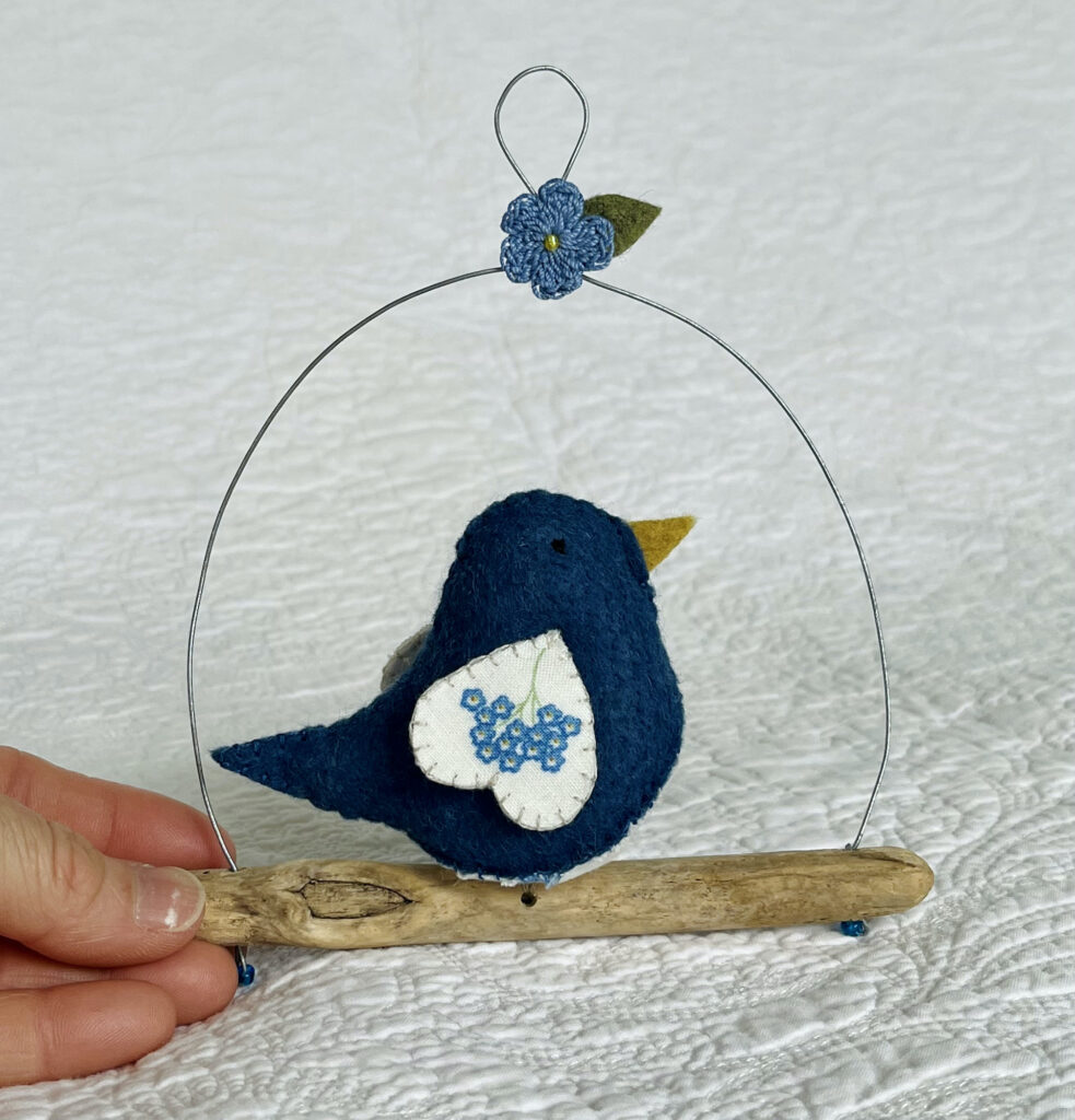 A single medium sized bird, handmade in denim blue felt with a cotton Forget-Me-Not flower print fabric chest and wings. The bird is sat on a natural driftwood perch with a wire hanger that is decorated with a crocheted               Forget-Me-Not flower and green leaf.