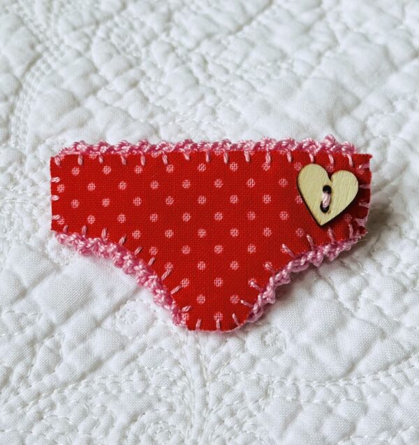 Handmade fabric and felt brooch with crocheted edging, a wooden heart button detail and a metal locking fastening. Approximate size 6cm Width x 4cm Height.