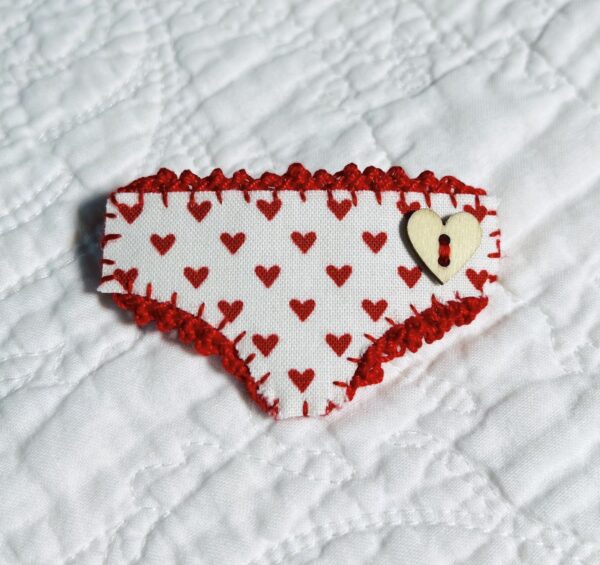 Handmade fabric and felt brooch with crocheted edging, a wooden heart button detail and a metal locking fastening. Approximate size 6cm Width x 4cm Height.