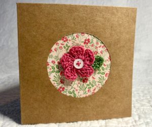 A greetings card with a floral circular centre cut out and a crocheted pink flower and green leaf embellishment with button detail.