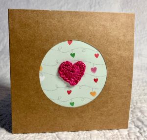 A small square, brown card greetings card with circular cut-out with patterned paper background and crocheted heart embellishment.