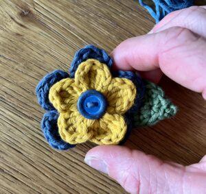 A mustard and denim blue flower made in 100% cotton, with a green leaf and blue wooden button detail.