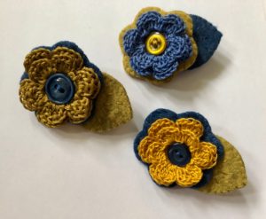 A crocheted cotton flower and embroidered felt leaf brooch with decorative button detail.