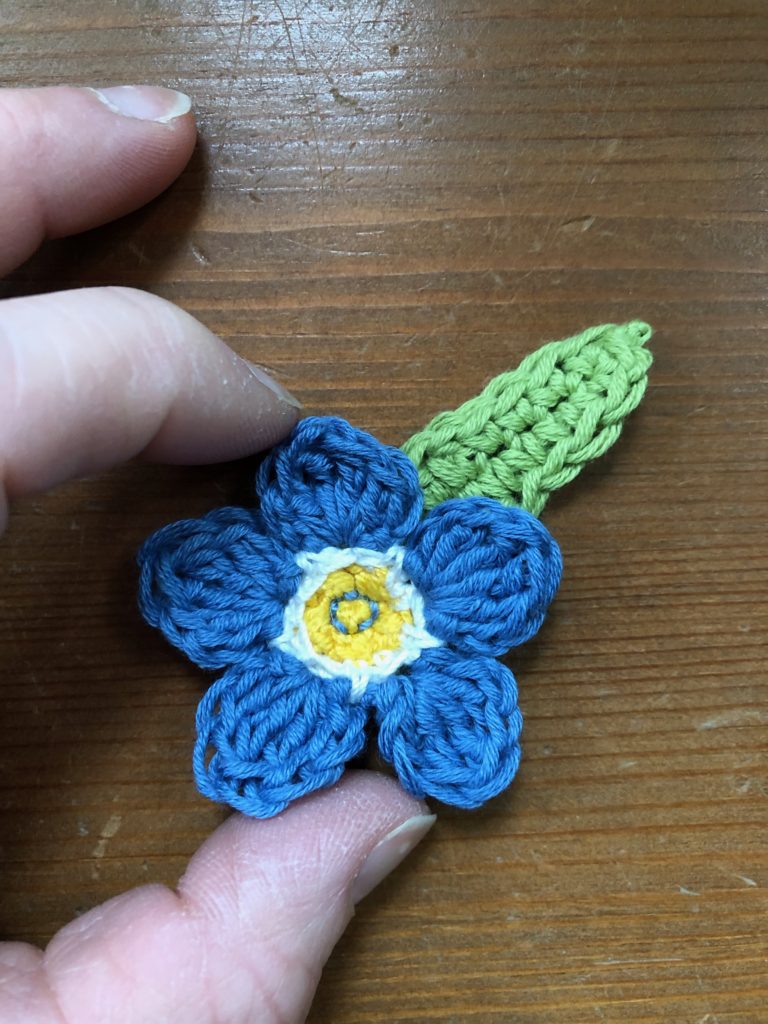A larger than life Forget-me-Not brooch, crocheted flower in purple with a green leaf.