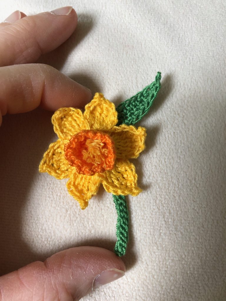 Crocheted yellow daffodil brooch with stem and green leaves.