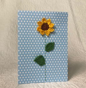 Greetings card. A blue spotty card with a crocheted sunflower and green crocheted leaves.