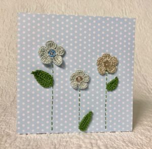 Greetings card. A blue spotty card with three blue and grey crocheted flowers and green crocheted leaves.