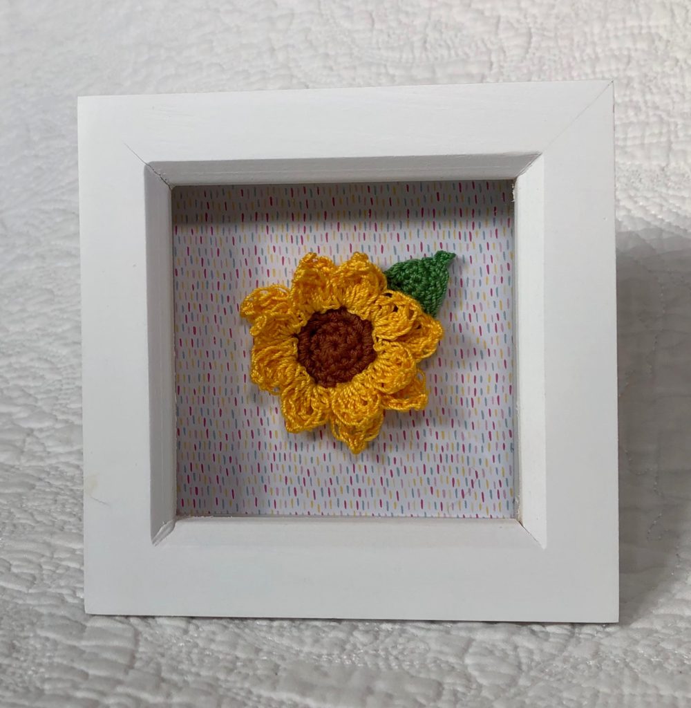 White wooden picture frame with small crocheted sunflower and leaf on a multi coloured paper background.