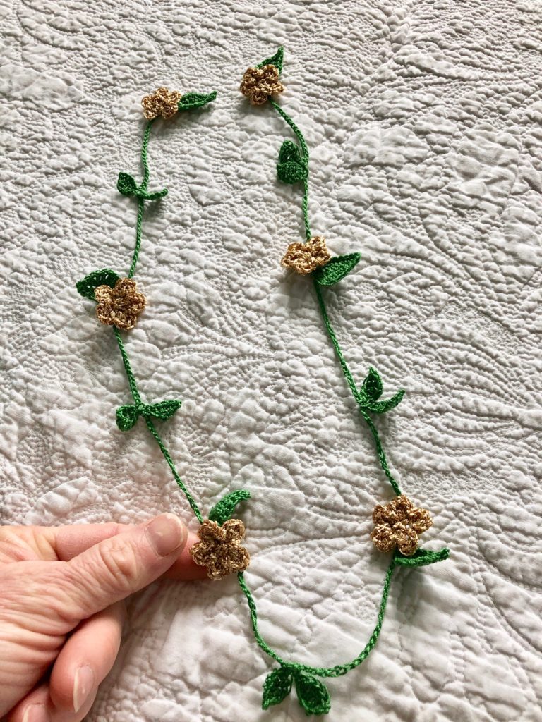 A headband garland of metallic golden crocheted flowers and golden glass beads on a garland of green crocheted leaves.