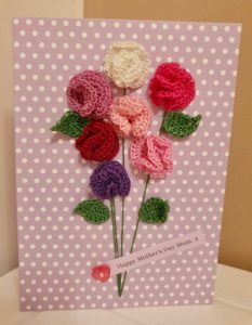 A greetings card with crocheted sweet peas in pinks and green leaves tied in a bunch with a label and button detail on a lilac spotty card.