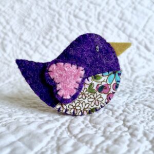 This little birdie brooch is completely hand cut, stitched, embroidered and embellished. It is made using a wool mix felt in dark purple and pink, with a floral patterned fabric chest. It has a metal locking fixing on the back.