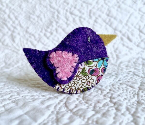 This little birdie brooch is completely hand cut, stitched, embroidered and embellished. It is made using a wool mix felt in dark purple and pink, with a floral patterned fabric chest. It has a metal locking fixing on the back.