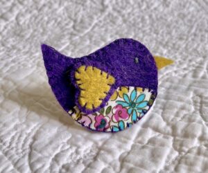 This little birdie brooch is completely hand cut, stitched, embroidered and embellished. It is made using a wool mix felt in dark purple and mustard colours with a floral patterned fabric chest. It has a metal locking fixing on the back.