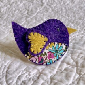 This little birdie brooch is completely hand cut, stitched, embroidered and embellished. It is made using a wool mix felt in dark purple and mustard colours with a floral patterned fabric chest. It has a metal locking fixing on the back.