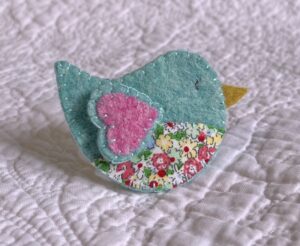 This little birdie brooch is completely hand cut, stitched, embroidered and embellished. It is made using a wool mix felt in light green and pink colours with a floral patterned fabric chest. It has a metal locking fixing on the back.