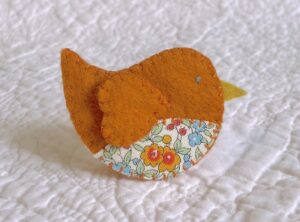 This little birdie brooch is completely hand cut, stitched, embroidered and embellished. It is made using a wool mix felt in rust orange colour with a floral patterned fabric chest. It has a metal locking fixing on the back.