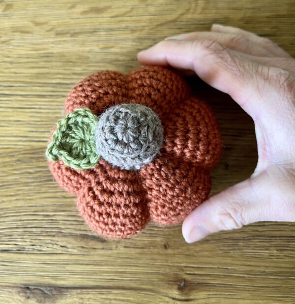 A medium sized, handmade, crocheted pumpkin made in a dark orange spice colour using 100% wool. With a brown stem and green leaf detail.