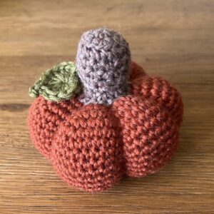 A medium sized, handmade, crocheted pumpkin made in a dark orange spice colour using 100% wool. With a brown stem and green leaf detail.