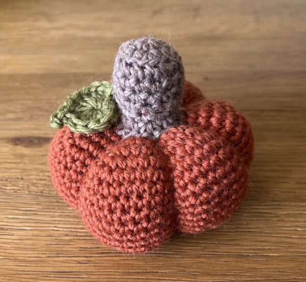 A medium sized, handmade, crocheted pumpkin made in a dark orange spice colour using 100% wool. With a brown stem and green leaf detail.