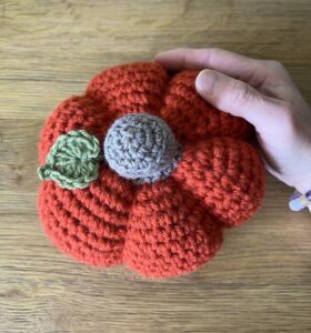 A medium sized, handmade, crocheted pumpkin made in a mix of autumnal colours. With a brown stem and green leaf detail.