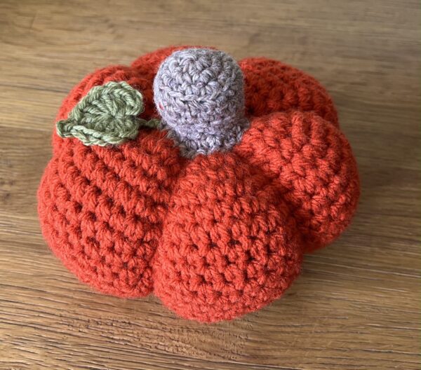 A large sized, handmade, crocheted pumpkin made in a spiced orange colour. With a brown stem and green leaf detail.