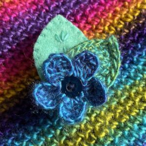 A single crocheted flower in a vibrant blue mix colour, with vintage button detail and two leaf brooch. Flower made using 100% acrylic with one crocheted leaf and one hand stitched felt leaf. A locking metal brooch fastening on the back. Approximate size 6cm width x 6cm height.