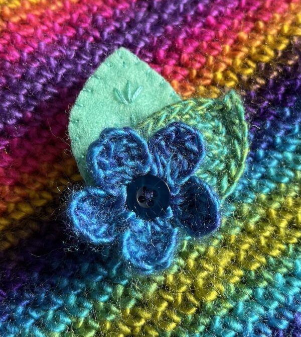 A single crocheted flower in a vibrant blue mix colour, with vintage button detail and two leaf brooch. Flower made using 100% acrylic with one crocheted leaf and one hand stitched felt leaf. A locking metal brooch fastening on the back. Approximate size 6cm width x 6cm height.