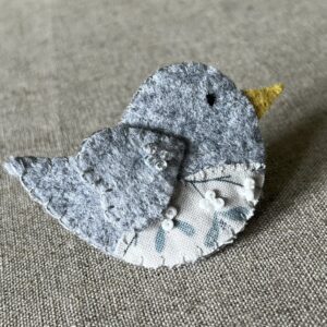 This little birdie brooch is completely hand cut, stitched, embroidered and embellished. It is made using a wool mix felt in a pale grey colour with a mistletoe patterned fabric chest. It is hand embellished using glass beads and has a metal locking fixing on the back.