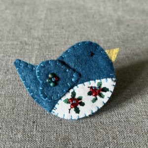 This little birdie brooch is completely hand cut, stitched, embroidered and embellished. It is made using a wool mix felt in a teal colour with a holly patterned fabric chest. It is hand embellished using glass beads and has a metal locking fixing on the back.