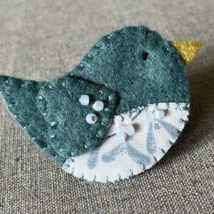 This little birdie brooch is completely hand cut, stitched, embroidered and embellished. It is made using a wool mix felt in a dark green colour with a mistletoe patterned fabric chest. It is hand embellished using glass beads and has a metal locking fixing on the back.