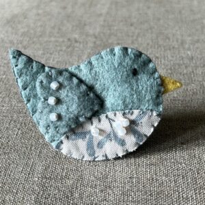 This little birdie brooch is completely hand cut, stitched, embroidered and embellished. It is made using a wool mix felt in a pale green colour with a mistletoe patterned fabric chest. It is hand embellished using glass beads and has a metal locking fixing on the back.