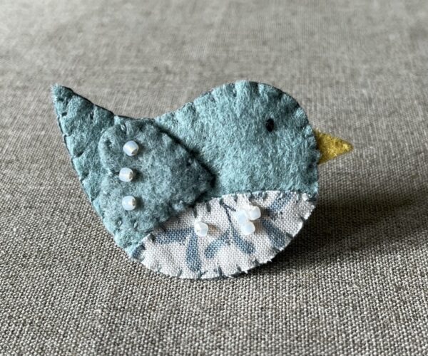 This little birdie brooch is completely hand cut, stitched, embroidered and embellished. It is made using a wool mix felt in a pale green colour with a mistletoe patterned fabric chest. It is hand embellished using glass beads and has a metal locking fixing on the back.