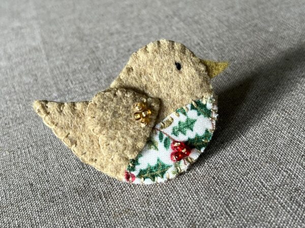 This little birdie brooch is completely hand cut, stitched, embroidered and embellished. It is made using a wool mix felt in a pale mustard colour with a holly patterned fabric chest. It is hand embellished using glass beads and has a metal locking fixing on the back.