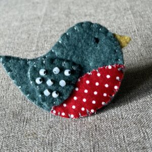 This little birdie brooch is completely hand cut, stitched, embroidered and embellished. It is made using a wool mix felt in a dark green colour with a spotty patterned fabric chest. It is hand embellished using glass beads and has a metal locking fixing on the back.