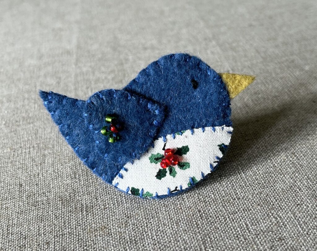 This little birdie brooch is completely hand cut, stitched, embroidered and embellished. It is made using a wool mix felt in a dark blue colour with a holly patterned fabric chest. It is hand embellished using glass beads and has a metal locking fixing on the back.