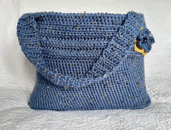 A handmade, crocheted shoulder bag with coordinating flower and leaf detail. Made in a denim blue, tweed style acrylic and wool mix yarn. Lined with a light blue heavy cotton fabric.