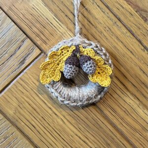 A handmade mini wreath hanging decoration. Crocheted in natural Jute and embellished with crocheted cotton oak leaves and acorns. Handmade using a wooden ring with 100% natural Jute, and 100% cotton embellishments. Approximate size of wreath 9cm height x 6.5cm width. Not including hanging loop.