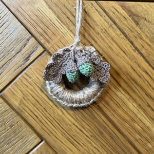 A handmade mini wreath hanging decoration. Crocheted in natural Jute and embellished with crocheted cotton oak leaves and acorns. Handmade using a wooden ring with 100% natural Jute, and 100% cotton embellishments. Approximate size of wreath 9cm height x 6.5cm width. Not including hanging loop.