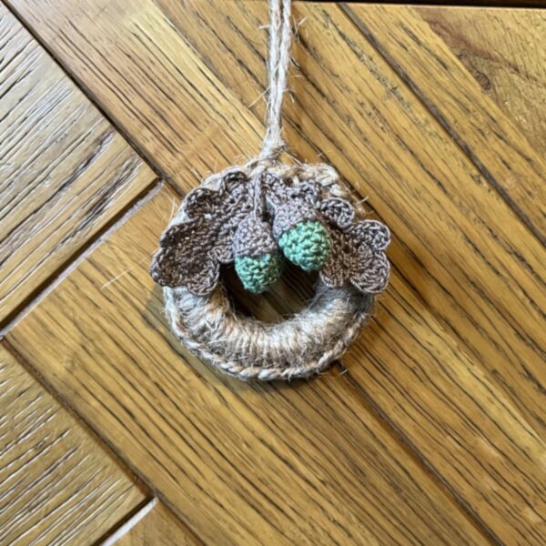 A handmade mini wreath hanging decoration. Crocheted in natural Jute and embellished with crocheted cotton oak leaves and acorns. Handmade using a wooden ring with 100% natural Jute, and 100% cotton embellishments. Approximate size of wreath 9cm height x 6.5cm width. Not including hanging loop.