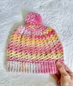 A soft and chunky textured hat with a large detachable bobble. Made in a subtle mix of pinks and yellows using a very soft and chunky, yet lightweight, 53% Wool/47% acrylic mix yarn. The bobble is made using a pink coloured 100% Acrylic yarn .