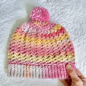 A soft and chunky textured hat with a large detachable bobble. Made in a subtle mix of pinks and yellows using a very soft and chunky, yet lightweight, 53% Wool/47% acrylic mix yarn. The bobble is made using a pink coloured 100% Acrylic yarn .