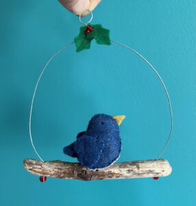 A single, small sized bird, handmade in denim blue felt with a cotton holly print fabric chest. The bird is sat on a natural driftwood perch with a wire hanger that is decorated with felt holly leaves and glass beads.