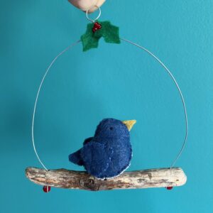 A single, small sized bird, handmade in denim blue felt with a cotton holly print fabric chest. The bird is sat on a natural driftwood perch with a wire hanger that is decorated with felt holly leaves and glass beads.